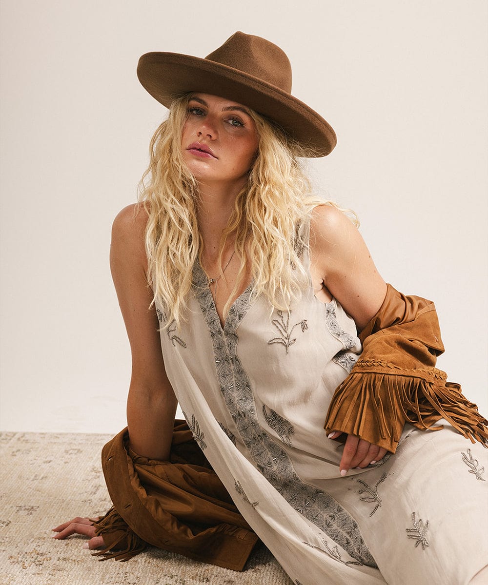 Woman wearing a chocolate colored pencil brim felt fedora hat and a white dress with floral patterns, sitting on a textured surface. #color_chocolate