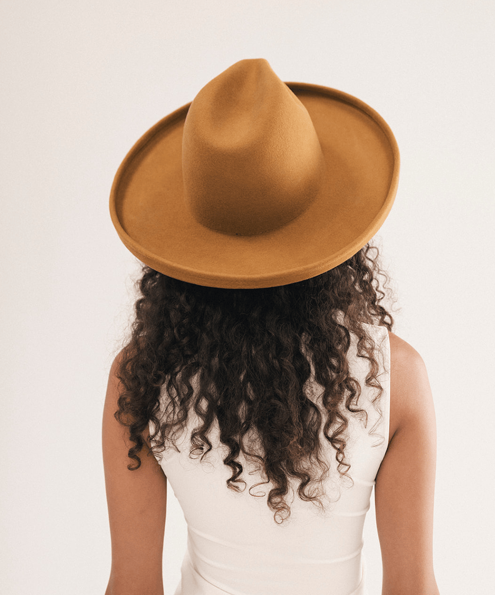 A person shown from the back wearing a wide-brimmed tan hat and a white sleeveless top, with long dark curly hair visible against a plain light background #color_cinnamon