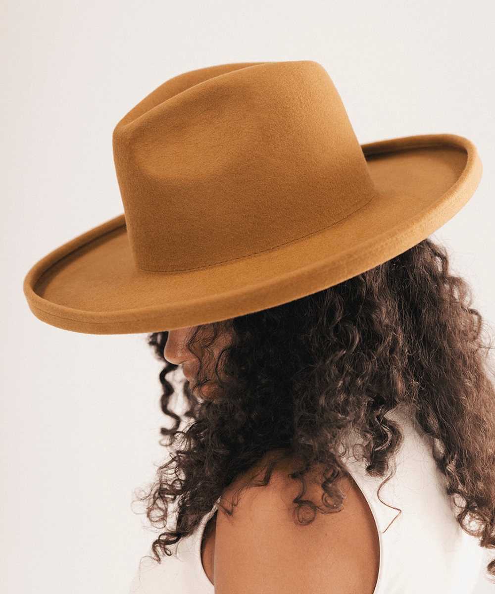 A woman with long, curly dark brown hair wearing a wide-brimmed felt hat in a warm camel brown color, photographed against a clean off-white background #color_cinnamon