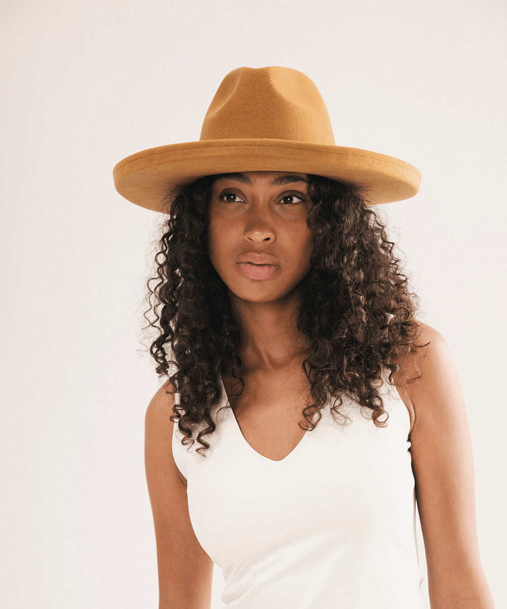 A woman with curly dark hair wearing a tan wide-brim fedora hat and a white sleeveless top, photographed against a plain light background #color_cinnamon
