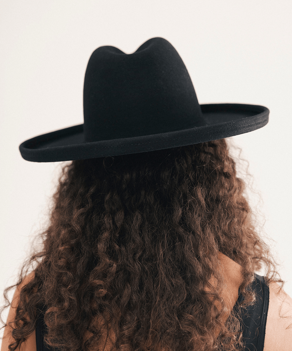 A person viewed from behind wearing a wide-brim black felt hat, with long curly brown hair and a black sleeveless top against a plain light background #color_black