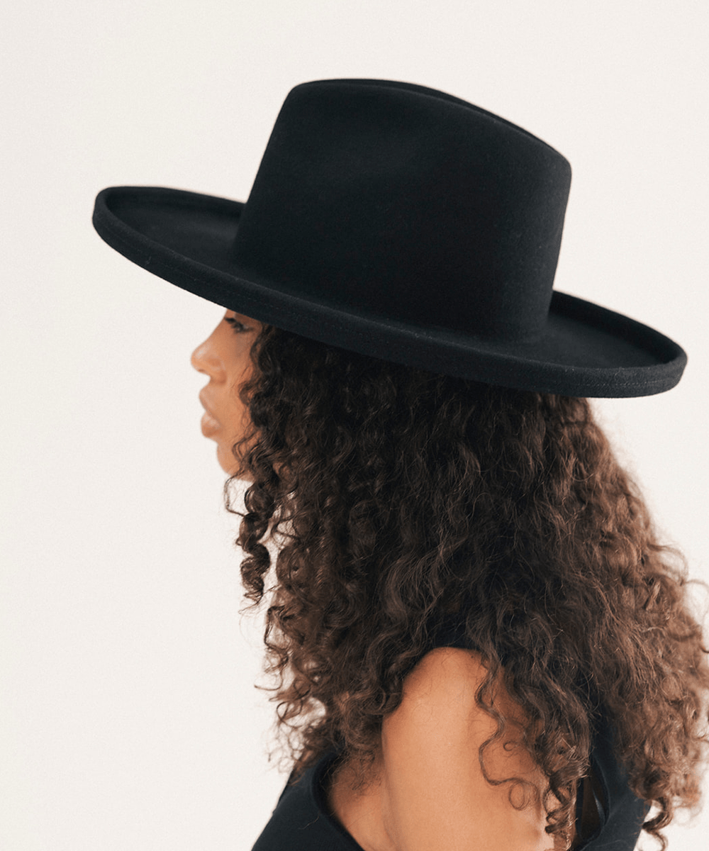A side-profile view of a woman with long, curly brown hair wearing a wide-brimmed black felt hat against a plain off-white background #color_black