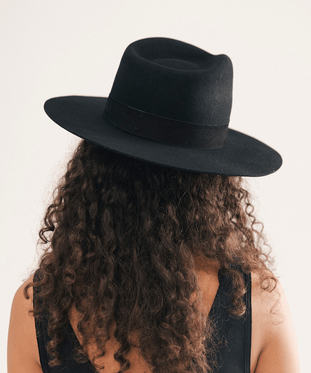 A person shown from the back wearing a wide-brim black felt hat, with long curly brown hair and a black sleeveless top, against a plain light background #color_black