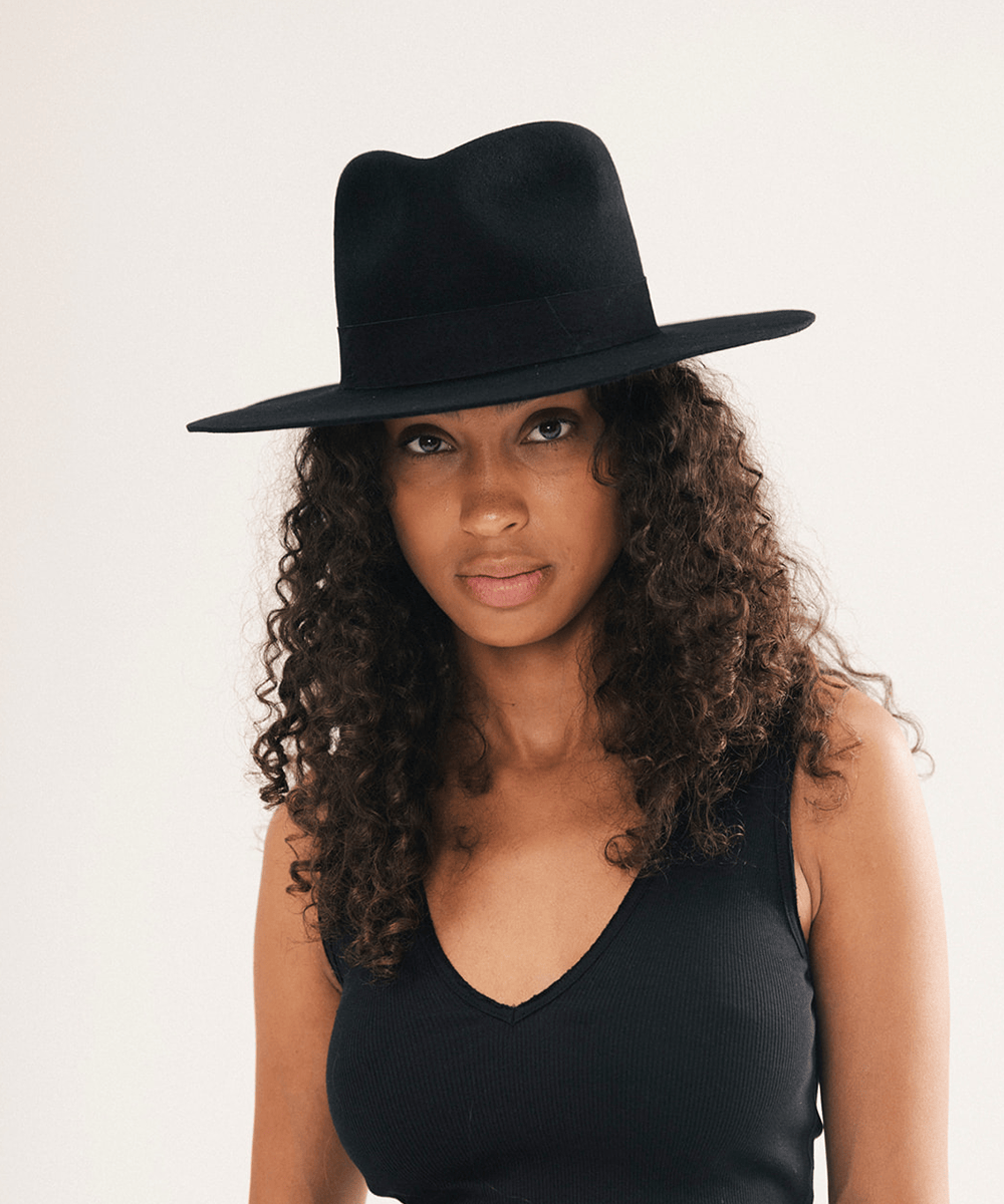 A woman with long, curly brown hair wearing a black wide-brim fedora hat and a black ribbed tank top, photographed against a plain off-white studio background #color_black
