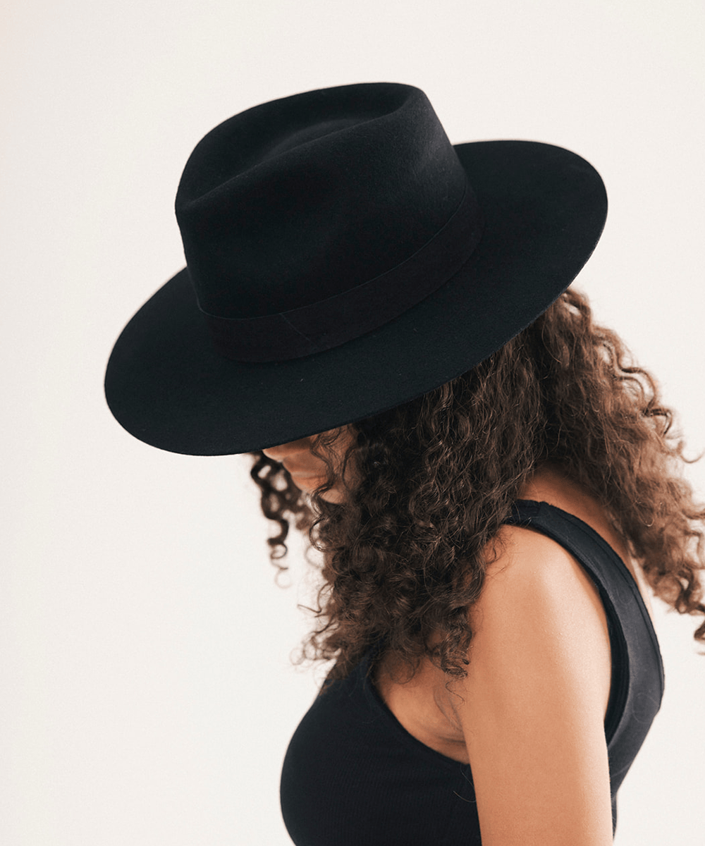A woman with curly brown hair wearing a black wide-brim fedora hat, shown in profile against a plain light background #color_black