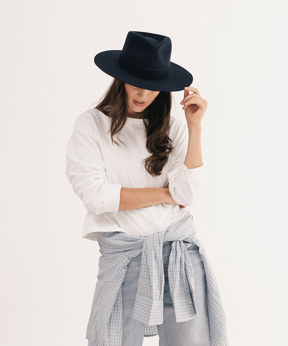 A woman wearing a black wide-brim fedora hat, a white long-sleeve top, and a light blue and white gingham shirt tied around her waist, posed against a plain white studio background #color_navy