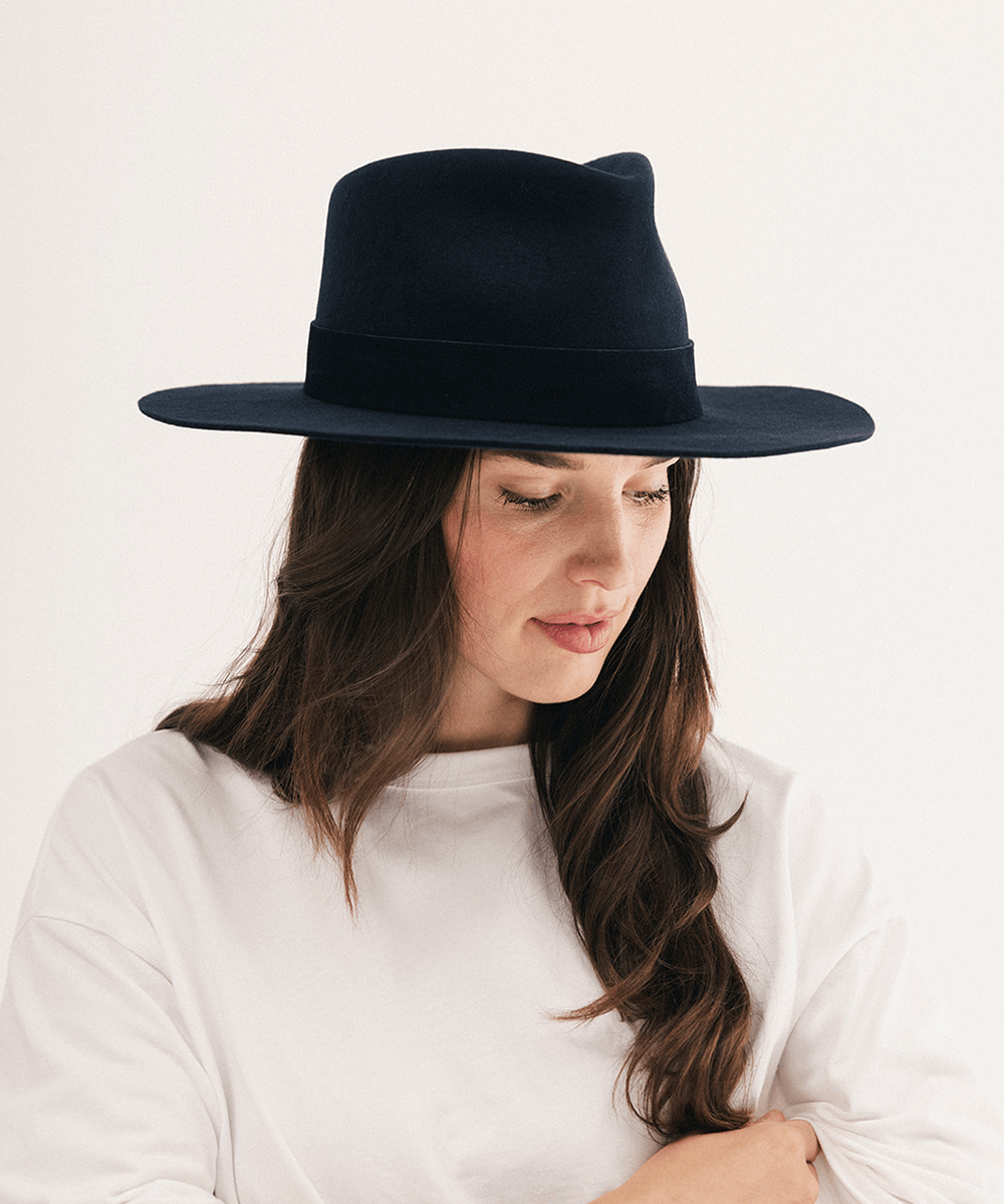 A woman wearing a wide-brimmed black felt fedora hat and a white long-sleeve top, photographed against a clean off-white studio background #color_navy