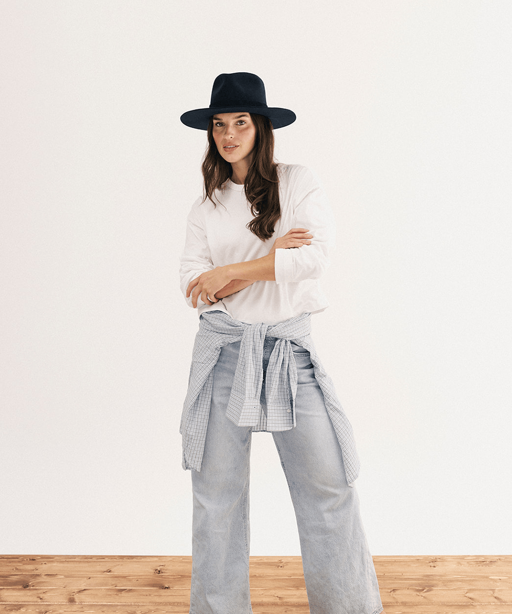A woman modeling a white long-sleeve shirt, light blue wide-leg jeans, a gray gingham shirt tied around her waist, and a black wide-brim hat against a white wall and wood floor #color_navy