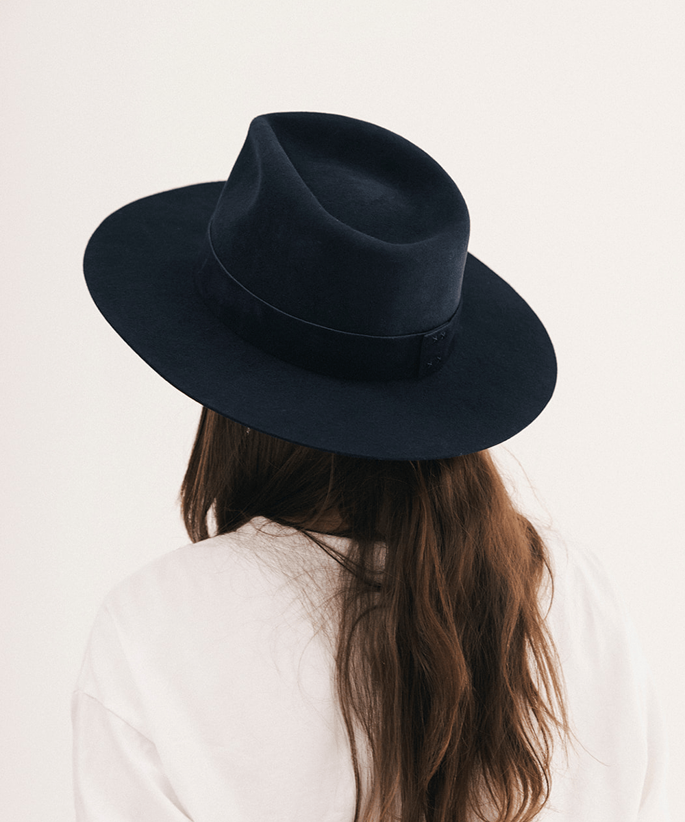 A person viewed from behind wearing a dark navy blue felt fedora hat with a matching band, paired with a white top against a plain off-white background #color_navy