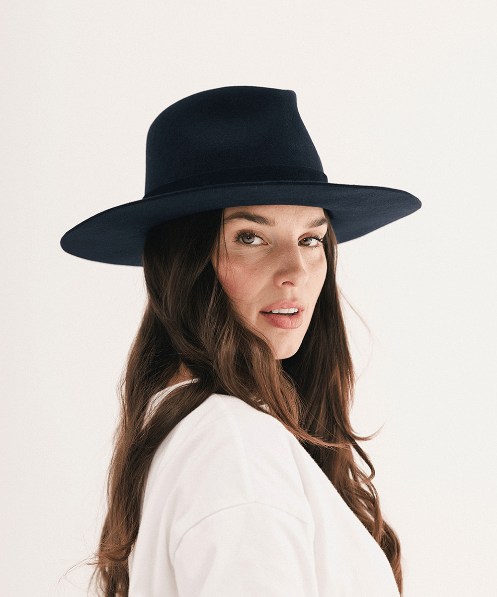 A woman with long brown hair wearing a black wide-brim fedora hat and a white top, photographed against a clean off-white studio background #color_navy