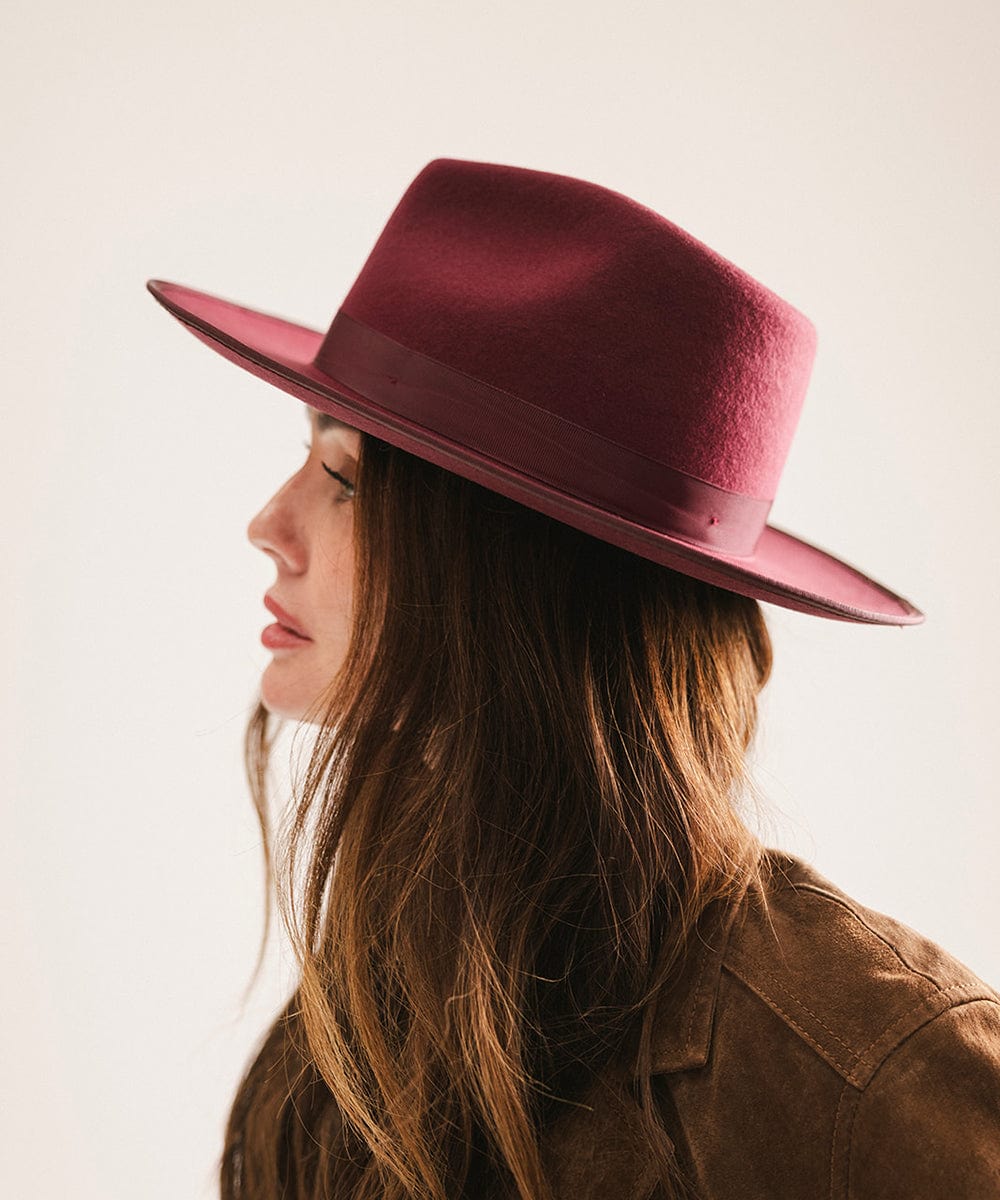 Woman wearing a merlot colored fedora hat with a band and a brown coat, facing sideways against a plain background #color_merlot
