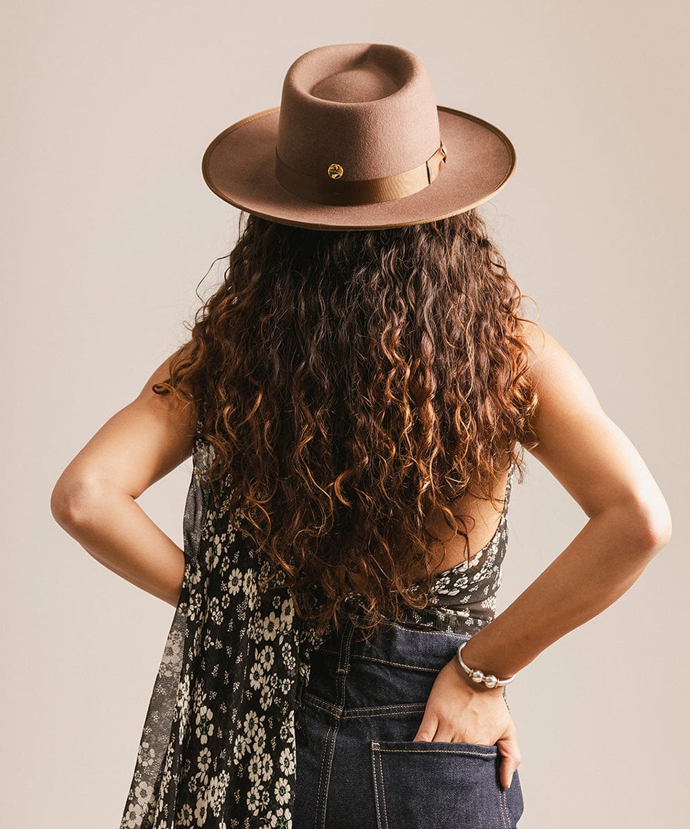 Woman wearing a chocolate colored hat with a band, floral top, and dark blue jeans, facing behind against a plain background #color_chocolate