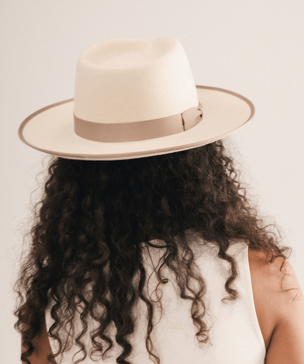 A person shown from behind wearing a cream-colored fedora hat with a taupe band and long, dark curly hair #color_white-taupe