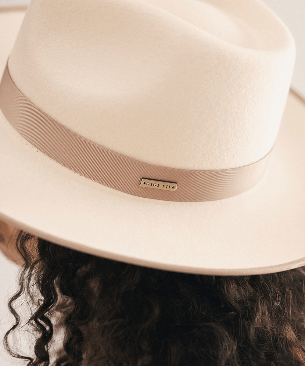 A close-up of a cream-colored felt fedora hat with a dusty pink ribbon band and a small gold-tone 'AUSLIE PIERP' metal plaque, worn by a model with dark curly hair #color_white-taupe