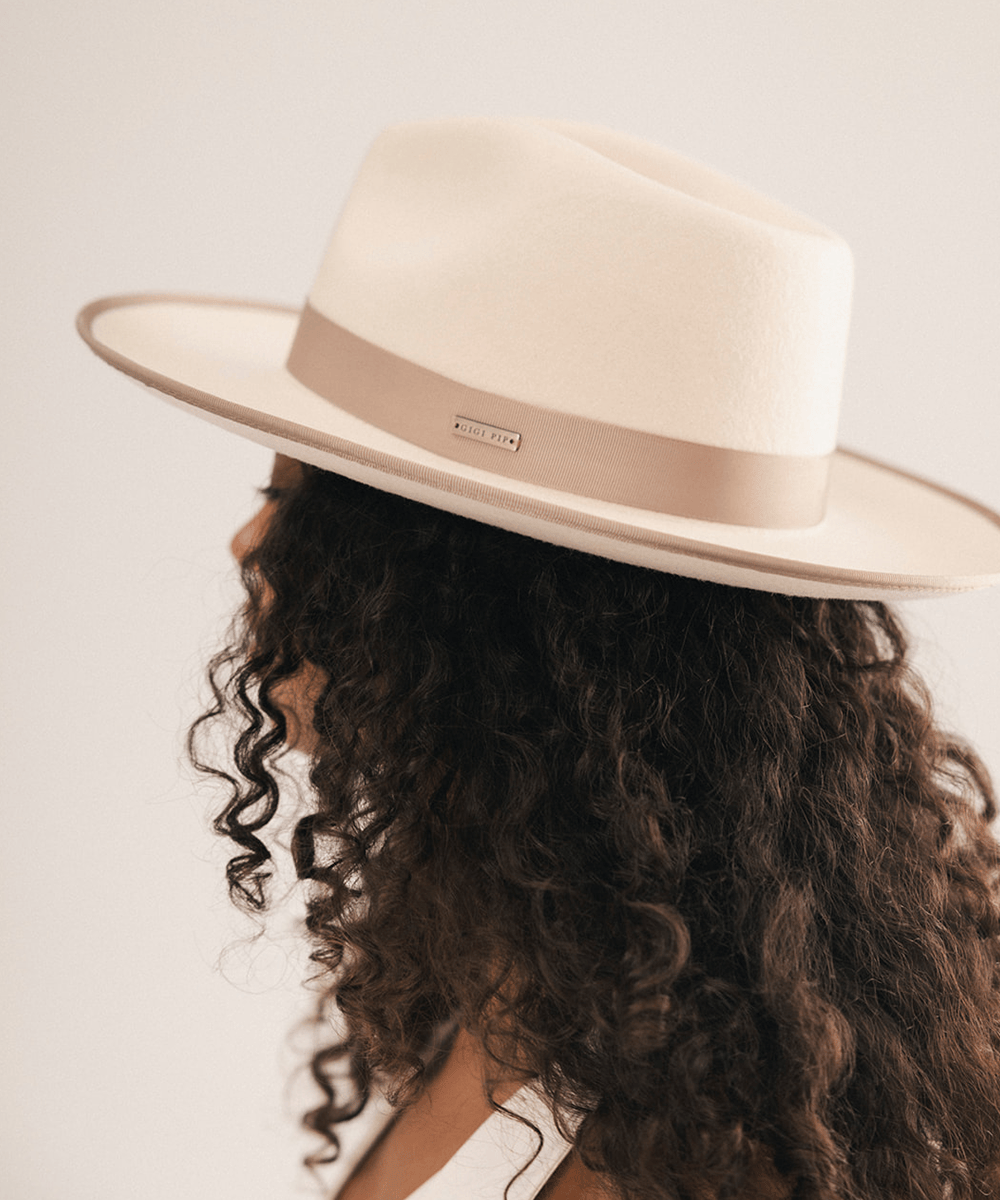 A woman shown from the side wearing a cream-colored fedora hat with a beige band and a small gold-tone brand plaque. The hat is photographed against a clean, light off-white background #color_white-taupe