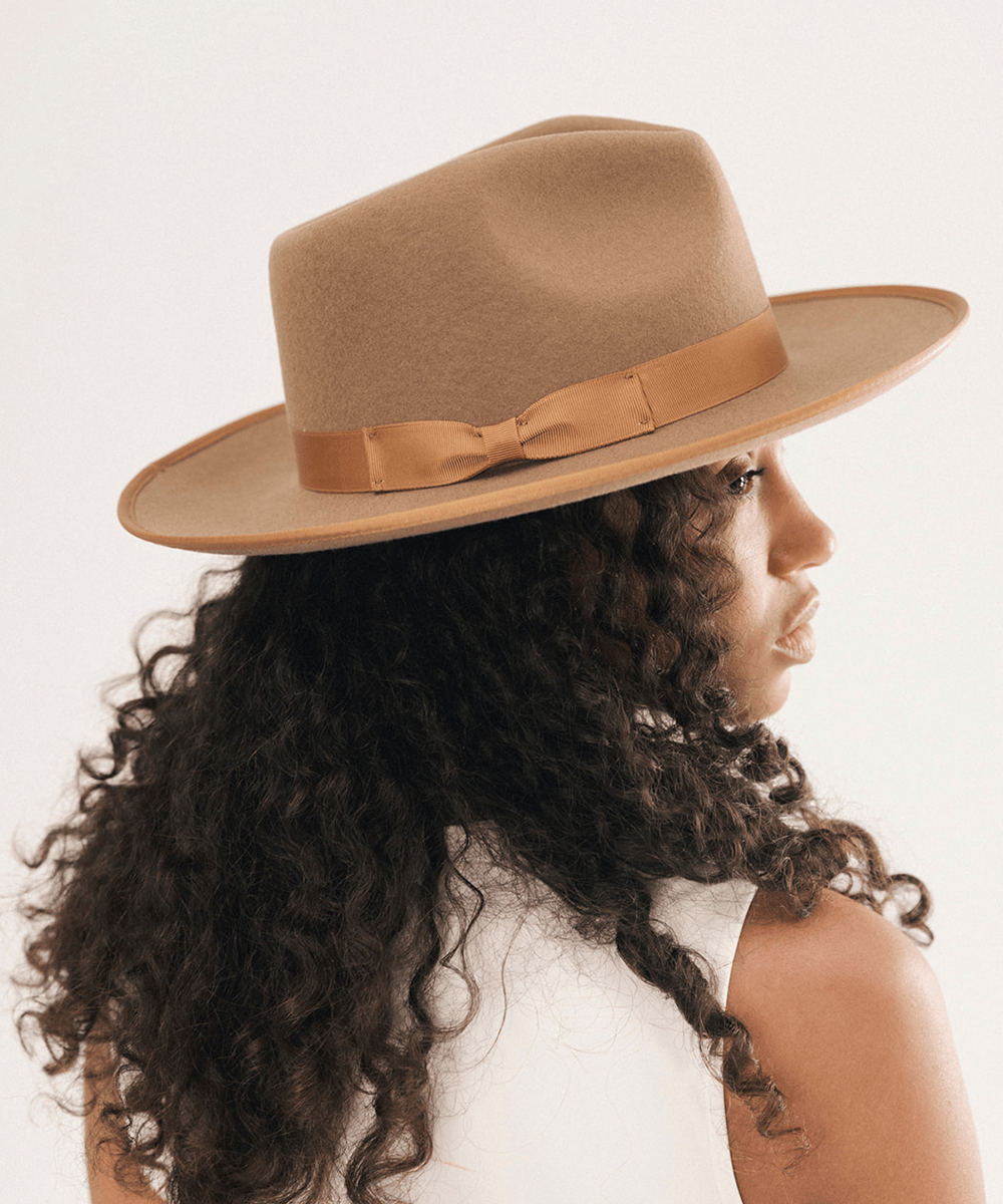 A woman with long, dark curly hair is shown from the back and side profile, wearing a wide-brim fedora hat in a warm camel brown with a matching brown ribbon band and bow. The background is a clean, off-white studio setting #color_brown