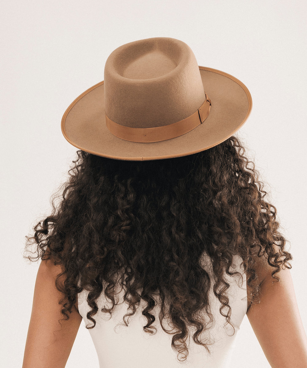 A person shown from the back wearing a wide-brim fedora hat in a warm camel tan color, with long dark curly hair and a white top against a plain light background #color_brown
