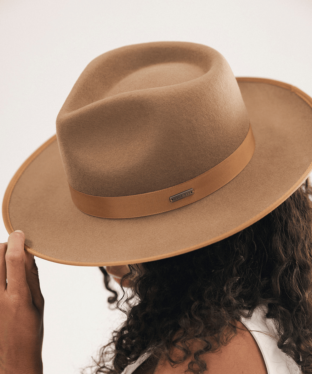 A close-up of a person wearing a tan felt fedora hat with a matching tan band and a small metal brand plaque, photographed against a plain light background #color_brown