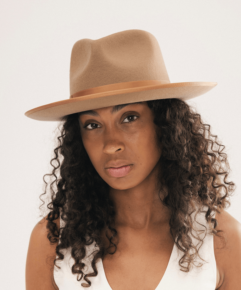 A woman with long, curly dark brown hair wearing a tan felt fedora hat with a matching band, photographed against a plain light background #color_brown