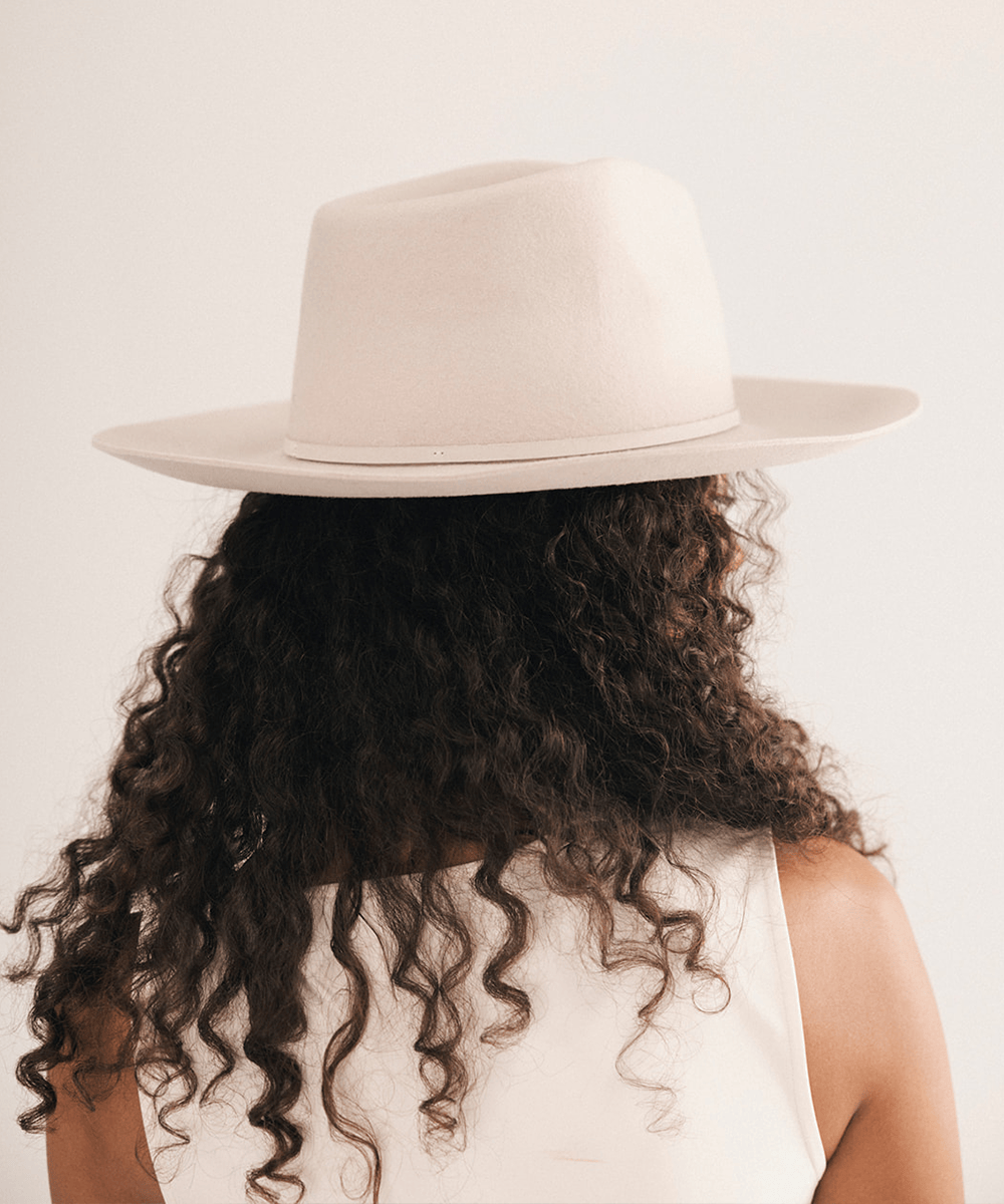 A person shown from the back wearing a wide-brim fedora hat and a white sleeveless top, with long dark curly hair, against a plain light background #color_ivory