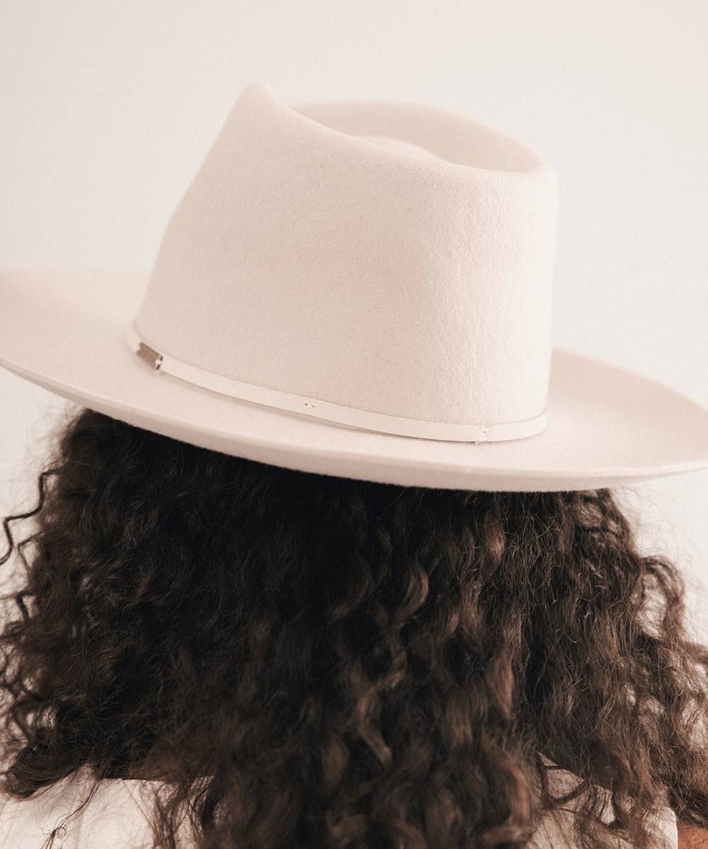 A close-up back view of a person wearing a wide-brim fedora hat. The hat is off-white/cream with a thin metallic band, and the person has dark curly hair #color_ivory