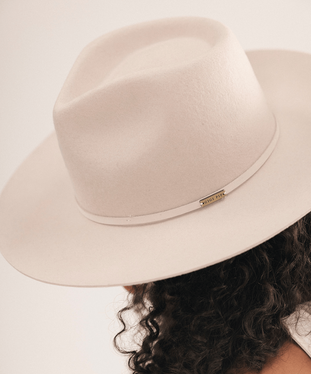 A close-up shot of a person wearing a light cream-colored felt fedora hat with a thin beige band and a small gold-tone metal brand plaque. The person has dark curly hair, and the background is a clean, solid off-white studio backdrop #color_ivory