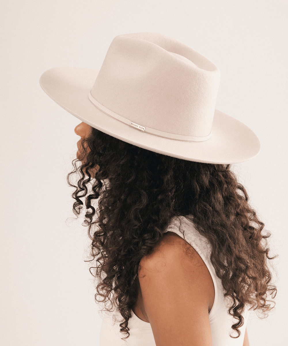 A woman shown in profile wearing a wide-brim fedora hat in a warm off-white or cream color, with long dark curly hair and a white sleeveless top against a plain light background #color_ivory