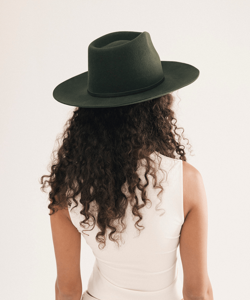 A woman viewed from behind wearing a dark olive green wide-brim fedora hat and a white sleeveless top, set against a plain light background #color_dark green
