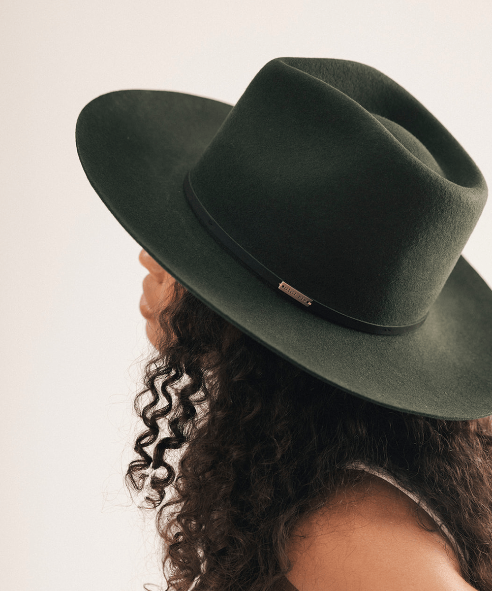 A dark olive green felt wide-brim hat with a black band and a small gold-tone metal accent, shown on a model with curly dark hair against a plain light background #color_dark green