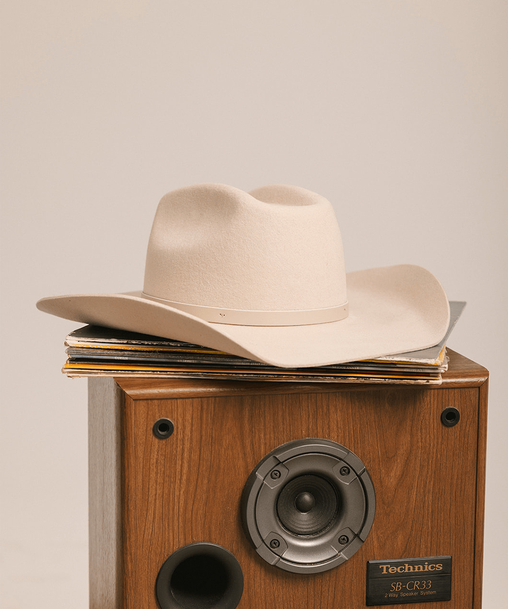 Cream cowboy hat with an upturned brim and an attached leather band on a wooden speaker against a plain background #color_cream