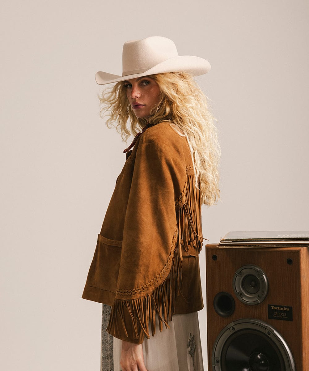 Woman wearing a cream cowboy hat with an upturned brim and an attached leather band, and a brown jacket standing next to a wooden speaker against a plain background #color_cream