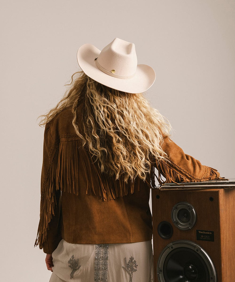 Woman wearing a cream cowboy hat with an upturned brim and an attached leather band, and a brown jacket, standing next to a wooden speaker and facing behind against a plain background #color_cream