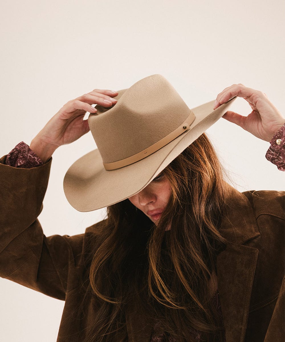 Woman wearing a tan cowboy hat with an upturned brim and an attached leather band and a brown suede jacket, looking down against a plain background #color_tan