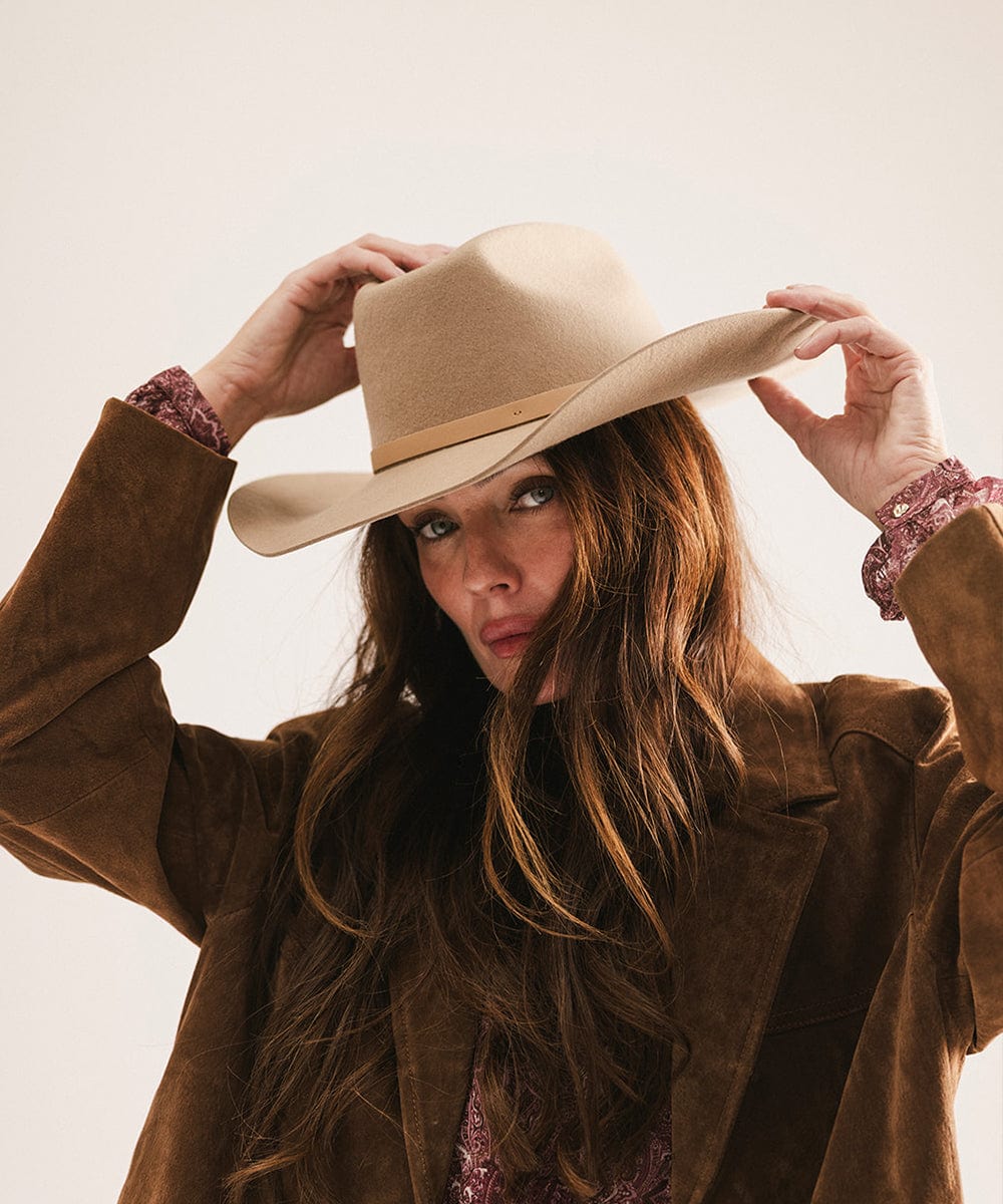 Woman wearing a tan cowboy hat with an upturned brim and an attached leather band and a brown suede jacket, posing against a plain background #color_tan