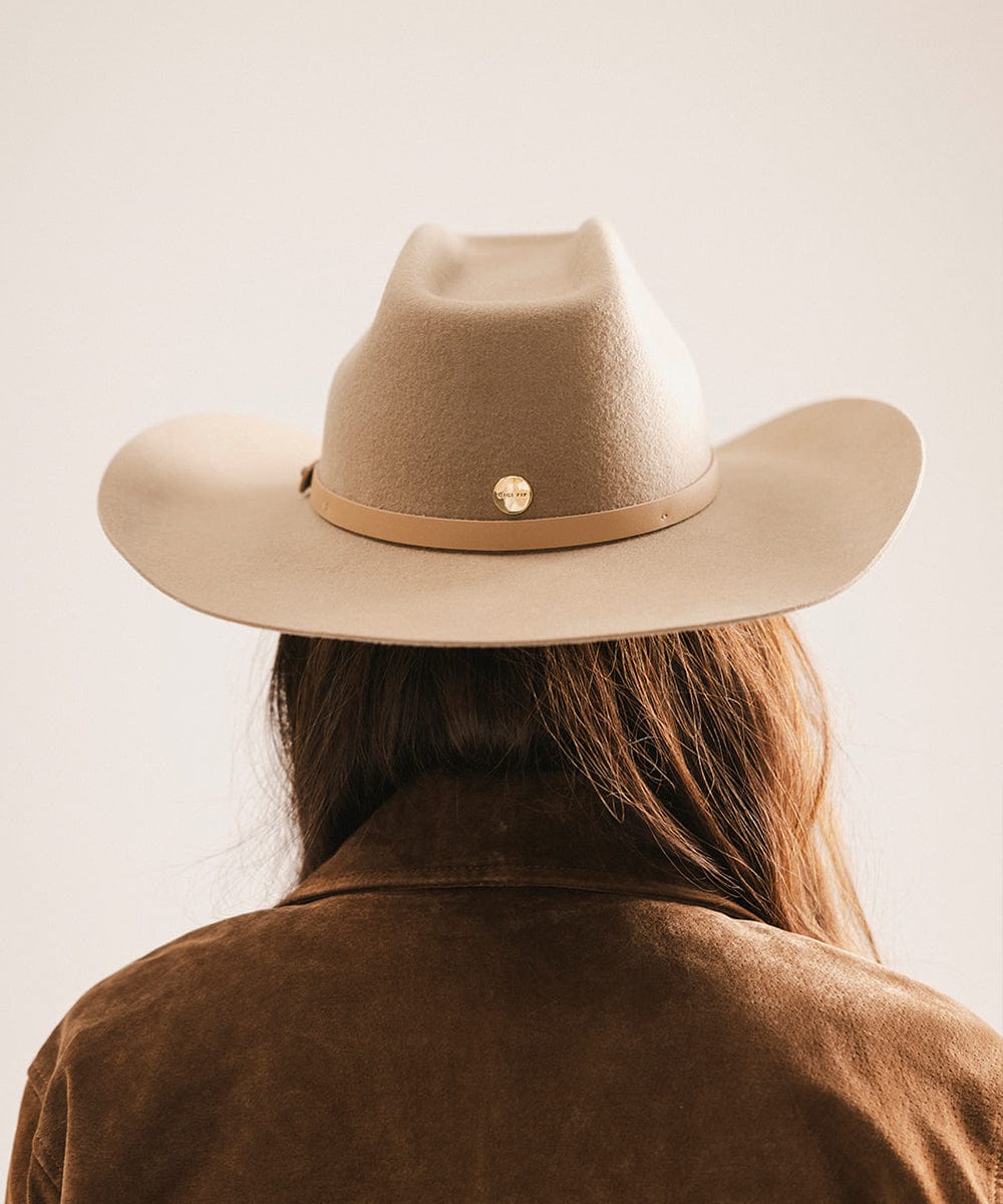 Woman wearing a tan cowboy hat with an upturned brim and an attached leather band and a brown suede jacket, facing behind against a plain background #color_tan