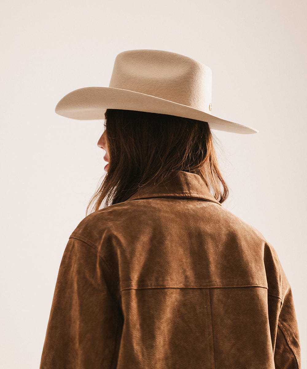 Woman wearing a tan cowboy hat with an upturned brim and an attached leather band and a brown suede jacket, facing sideways against a plain background #color_tan