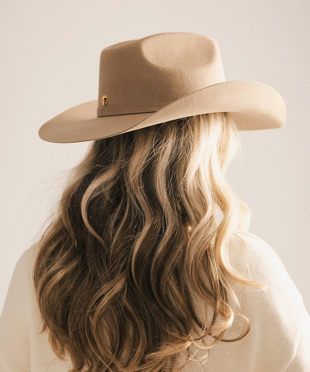 A woman shown from behind wearing a beige wide-brim cowboy hat with a small gold pin accent, styled with long wavy blonde hair and a cream knit top against a plain light background #color_tan