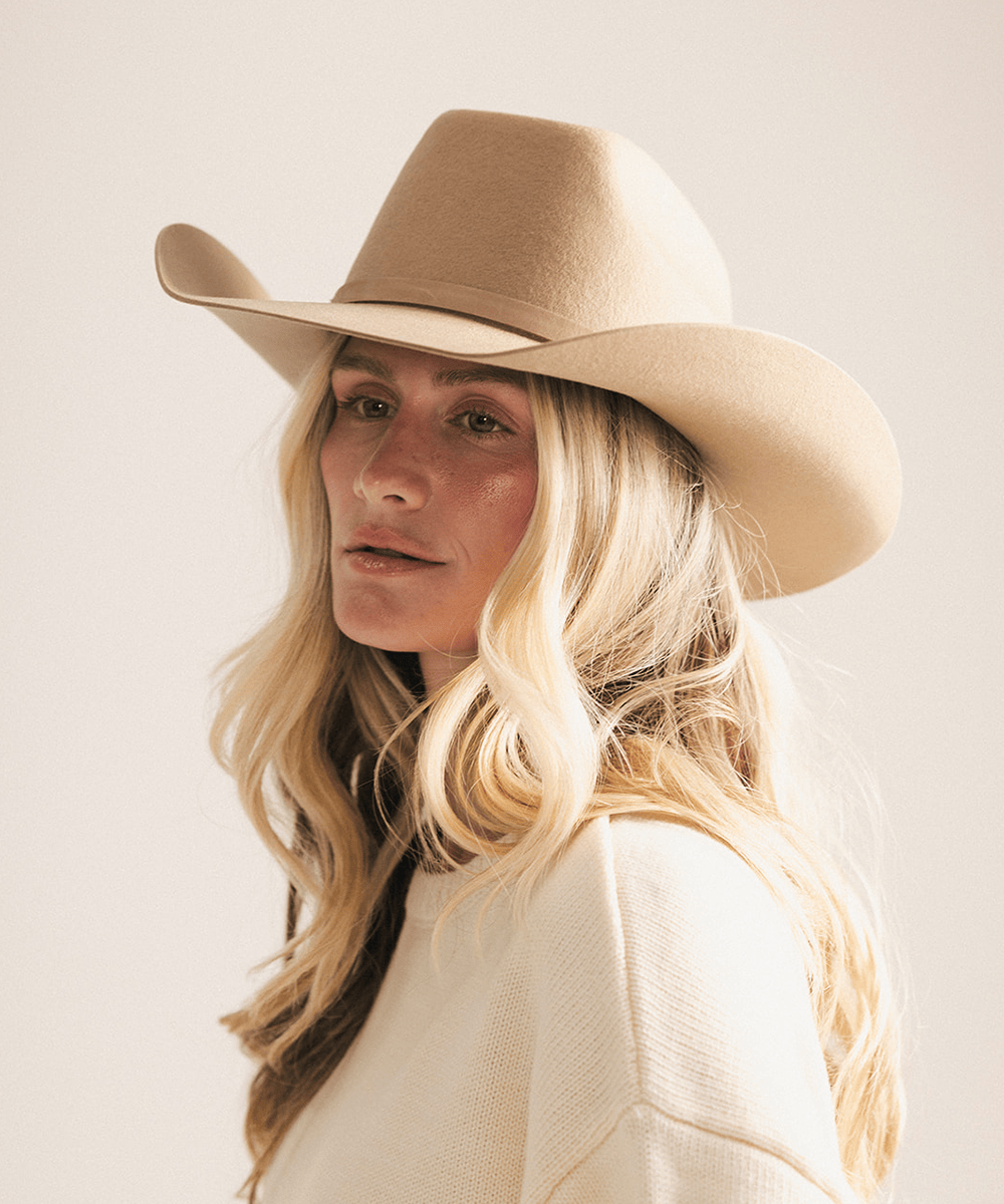 A blonde woman wearing a wide-brimmed beige cowboy hat and a cream knit sweater, photographed against a plain light beige background #color_tan