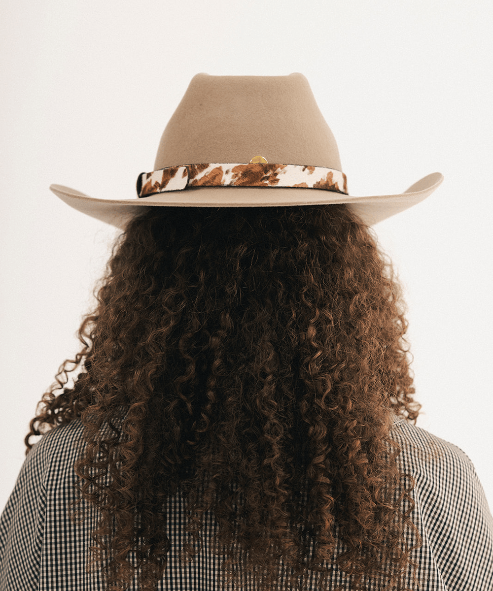 Woman wearing a tan cowboy hat with an upturned brim and an attached cow leather band, displayed against a white background #color_tan