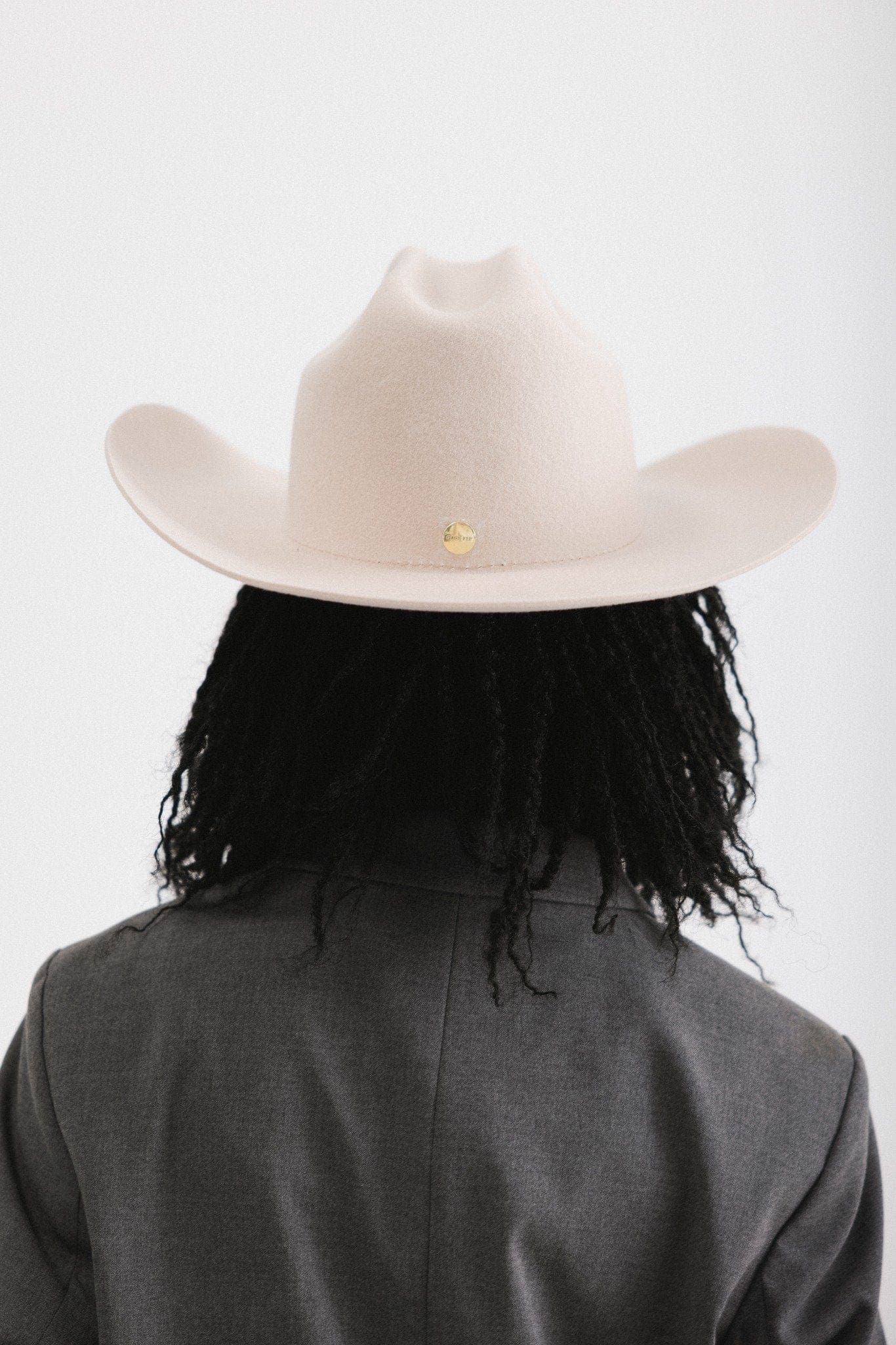 Woman wearing a cream cowboy hat and grey blazer, facing behind against a white background #color_cream