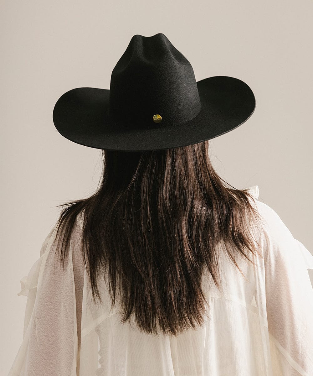 Woman wearing a black cowboy hat and a white shirt, facing behind against a plain background #color_black