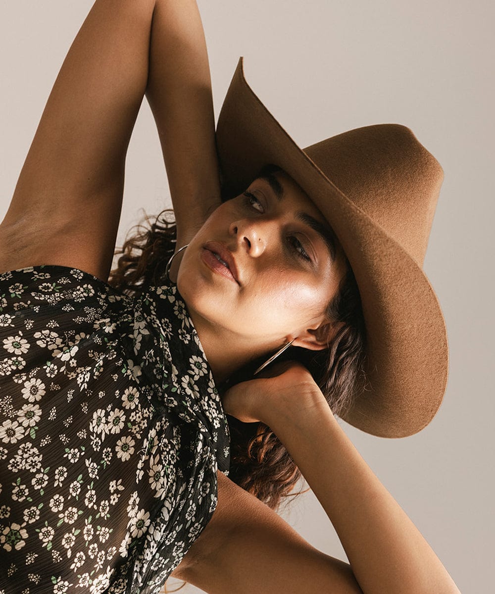 Woman wearing a caramel colored cowboy hat and black floral halter top, posing against a plain background #color_caramel