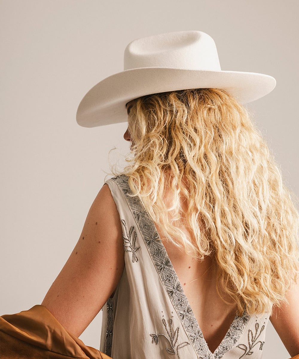Woman wearing an off white cowboy hat and a cream top, facing behind against a plain background #color_off white