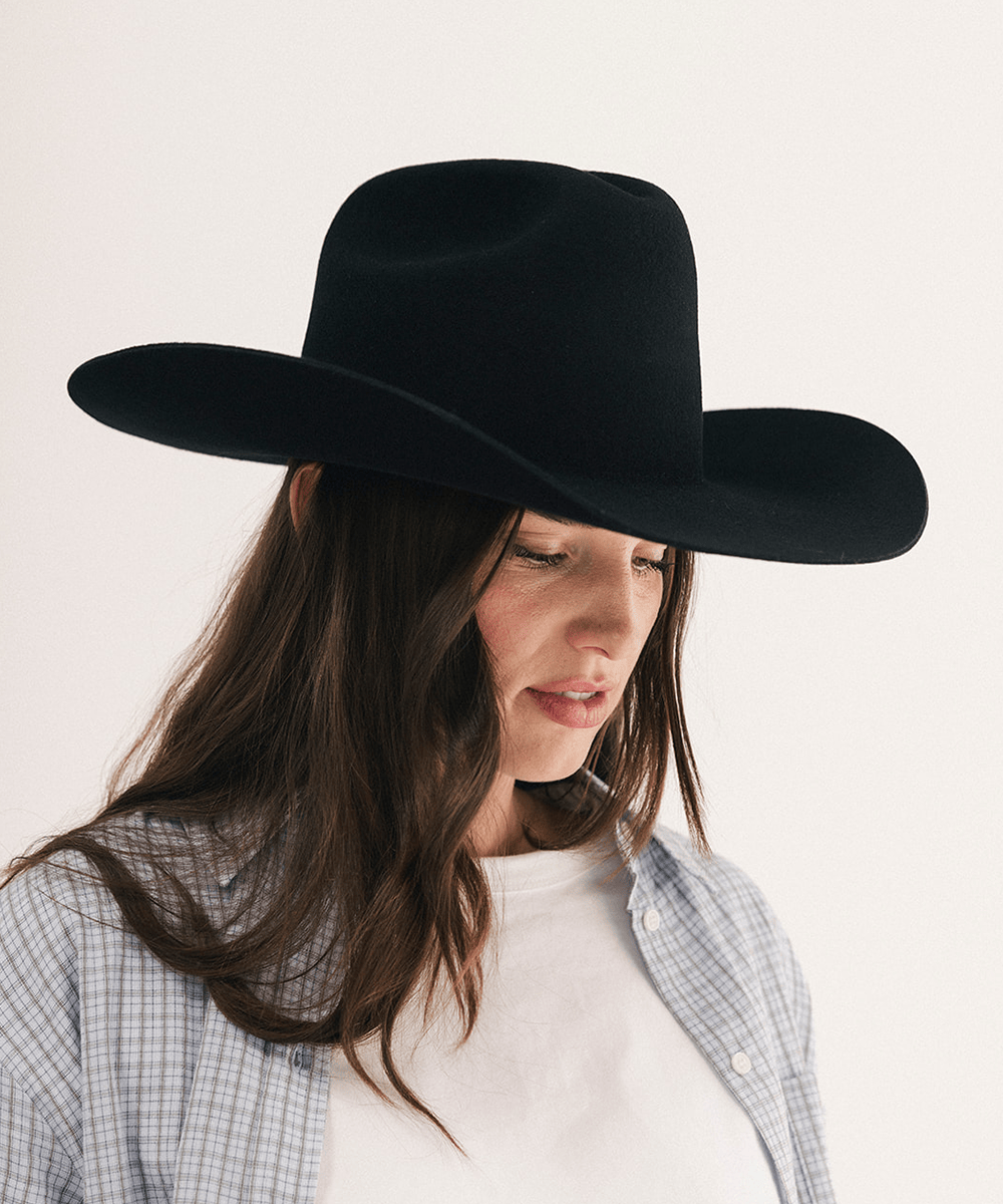 A woman wearing a wide-brim black cowboy hat, a light blue and white plaid button-up shirt, and a white t-shirt, photographed against a clean off-white background #color_black