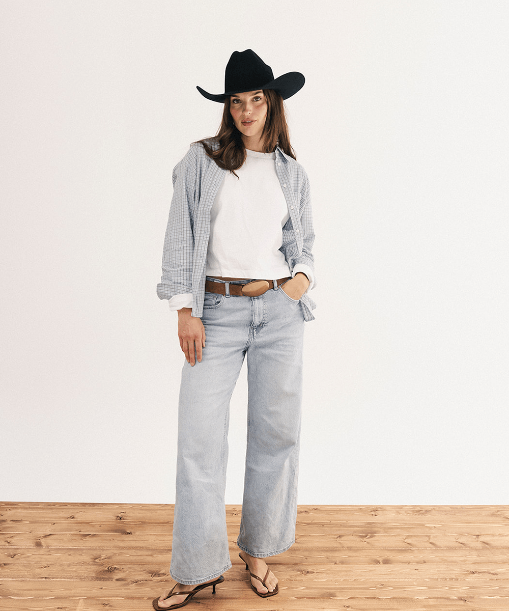 A woman stands in a studio wearing a black cowboy hat, a white crewneck tee, a light blue-and-white gingham button-up shirt, light-wash wide-leg jeans, a brown belt, and brown heeled sandals against a white wall and wood floor #color_black