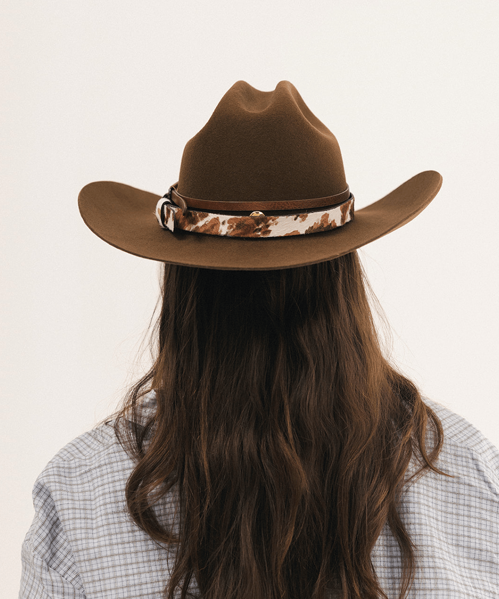 A person viewed from behind wearing a brown cowboy hat with a brown and white cow-print band, against a plain light background #color_coffee