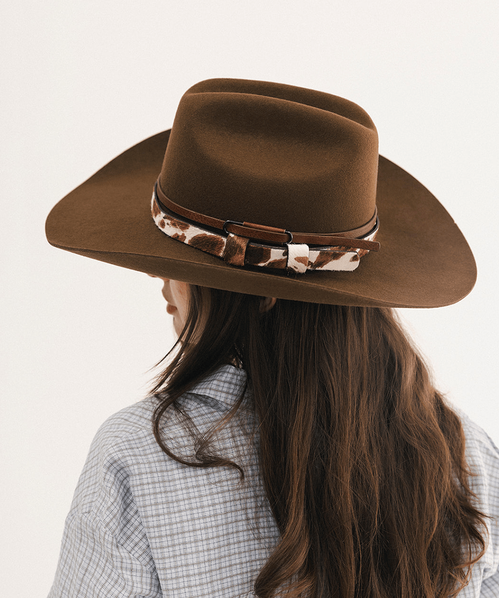 A woman shown from the back wearing a brown wide-brim felt cowboy hat with a brown leather band and a brown-and-white cowhide-patterned hatband, against a plain off-white background #color_coffee