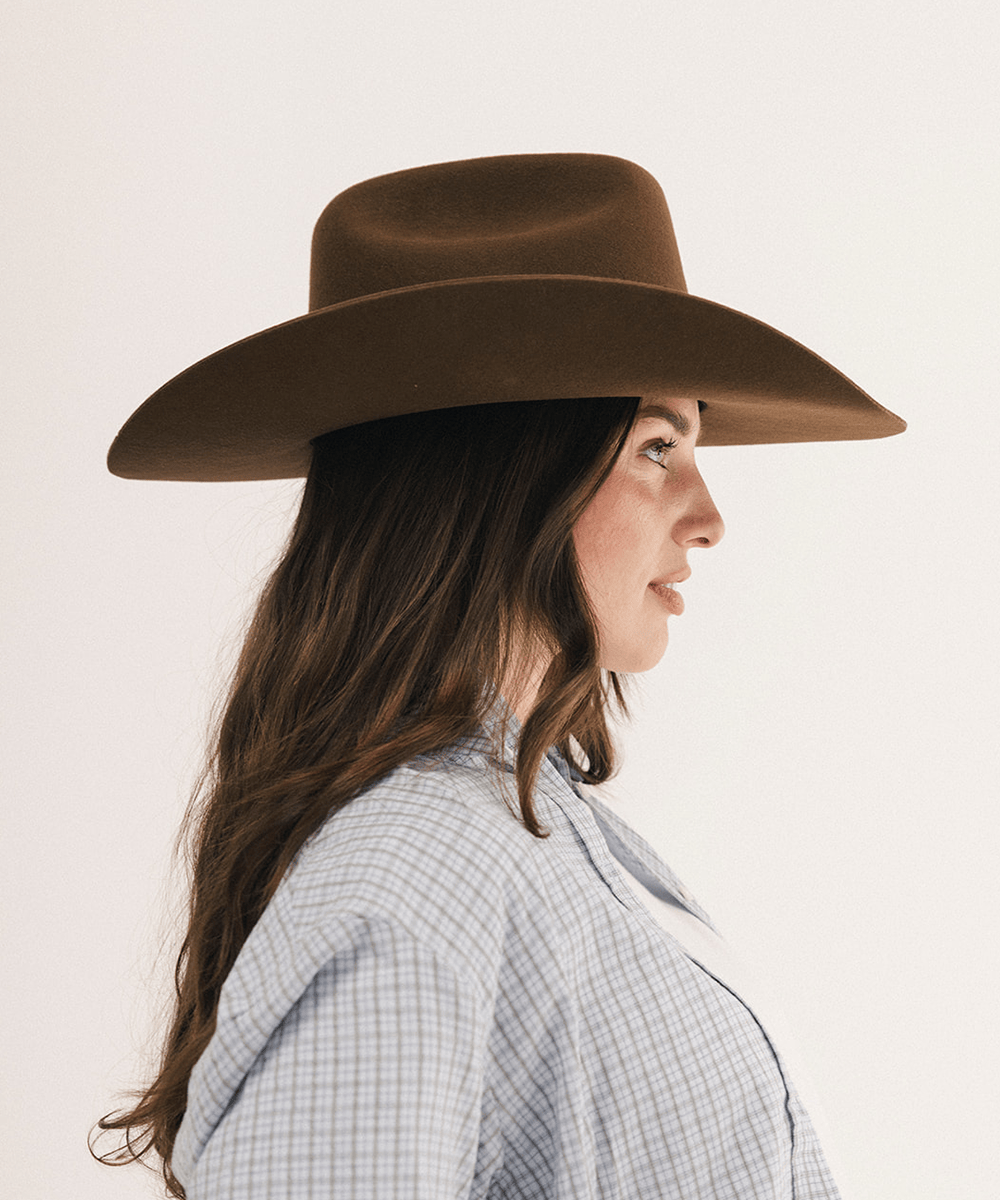A woman in side profile wearing a wide-brimmed brown felt hat and a light blue and white checkered shirt, set against a plain off-white background #color_coffee