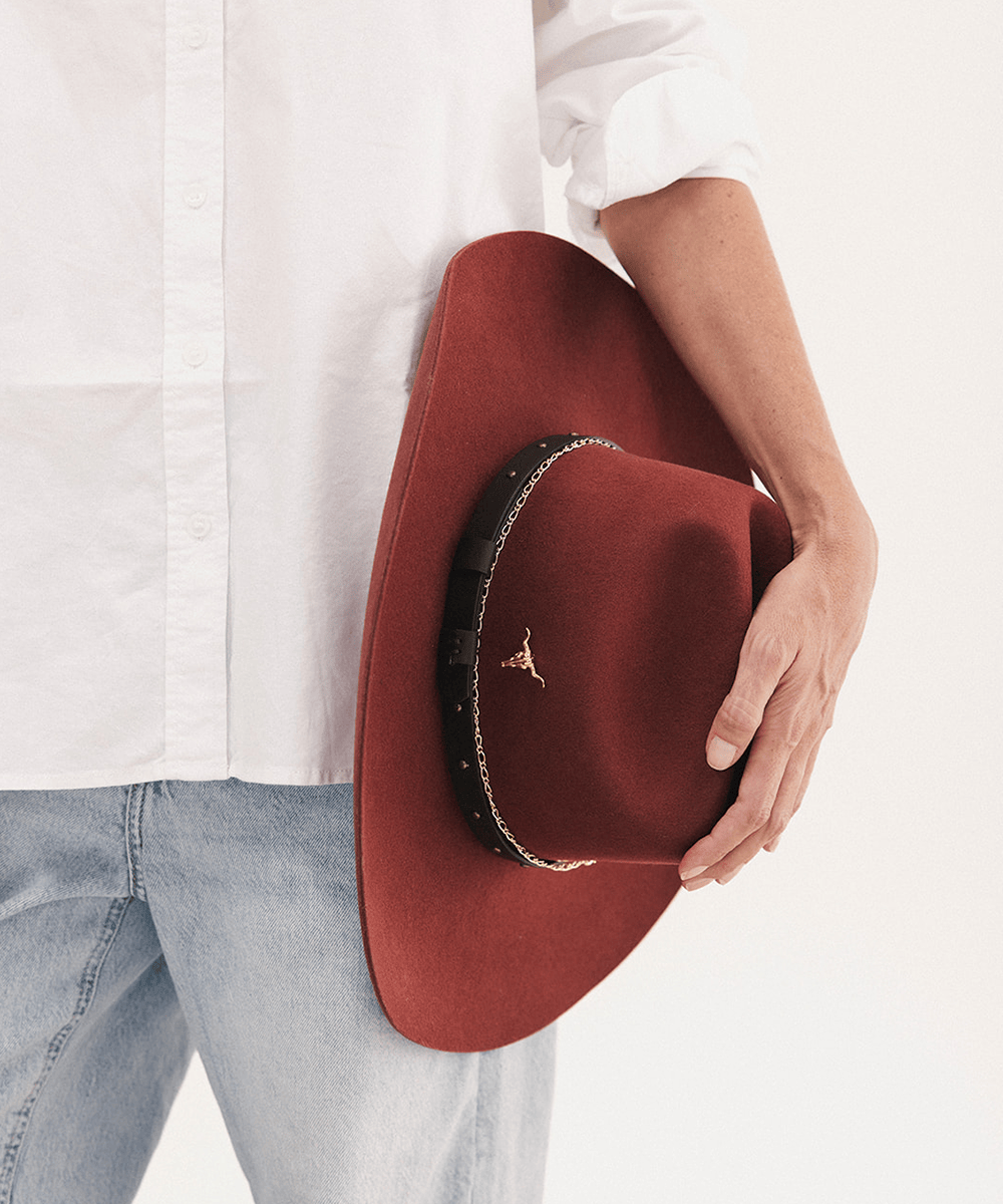 A person in a white button-up shirt and light blue jeans holds a wide-brimmed felt hat in rust red. The hat features a black band with gold trim and a small gold logo on the front #color_rusty red