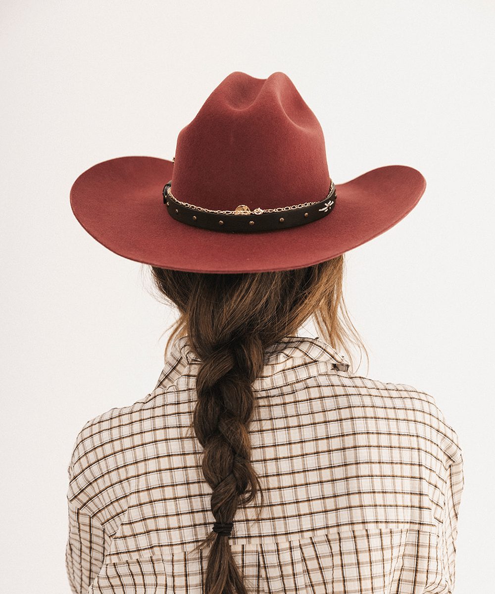 A person shown from the back wearing a wide-brimmed rust red cowboy hat with a black band and gold studs, paired with a beige and brown plaid shirt against a plain white background #color_rusty red